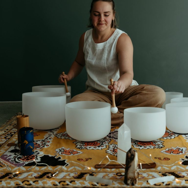 A woman sitting on a patterned rug playing white crystal singing bowls during a sound healing session.