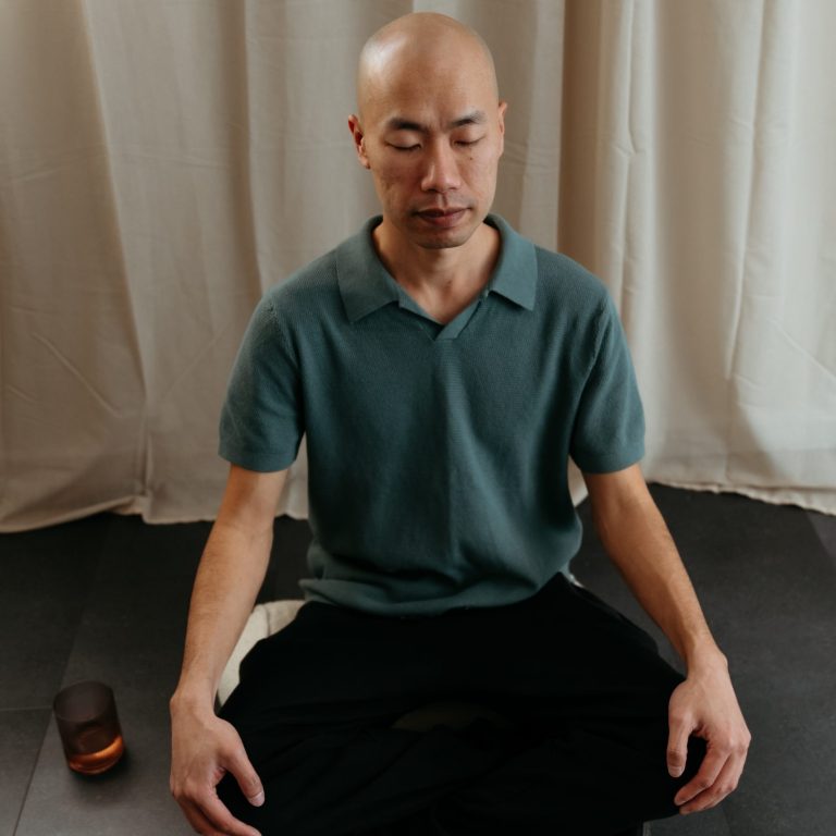 A person sitting cross-legged on the floor in meditation, wearing a green short-sleeved shirt and black pants, with eyes closed and a calm expression. A candle is placed beside them, and a light curtain hangs in the background