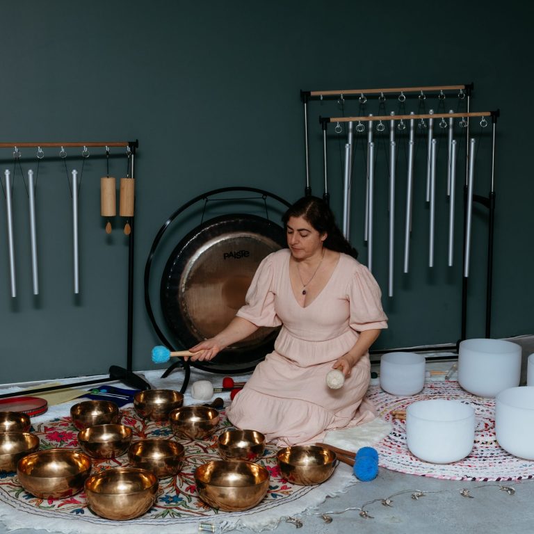 Alessandra seated on a rug surrounded by singing bowls, gongs, and chimes, gently playing during a sound healing session.
